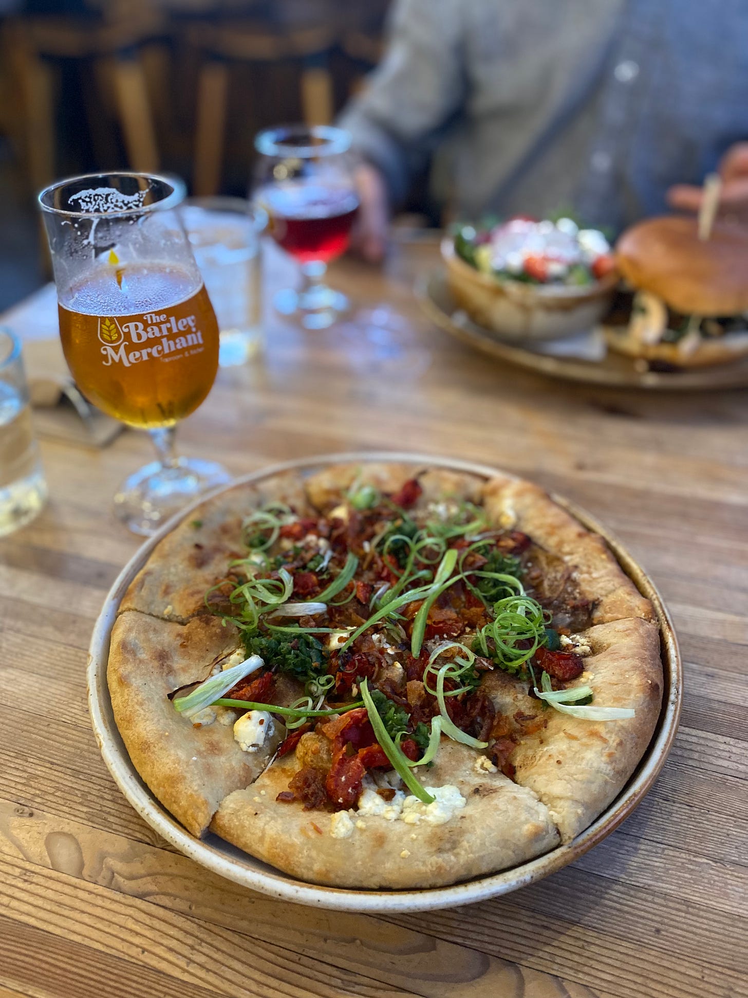 The flatbread described above, on a round ceramic plate and covered with swirls of thinly sliced green onion. Next to it is a half-empty beer in a branded tulip glass.