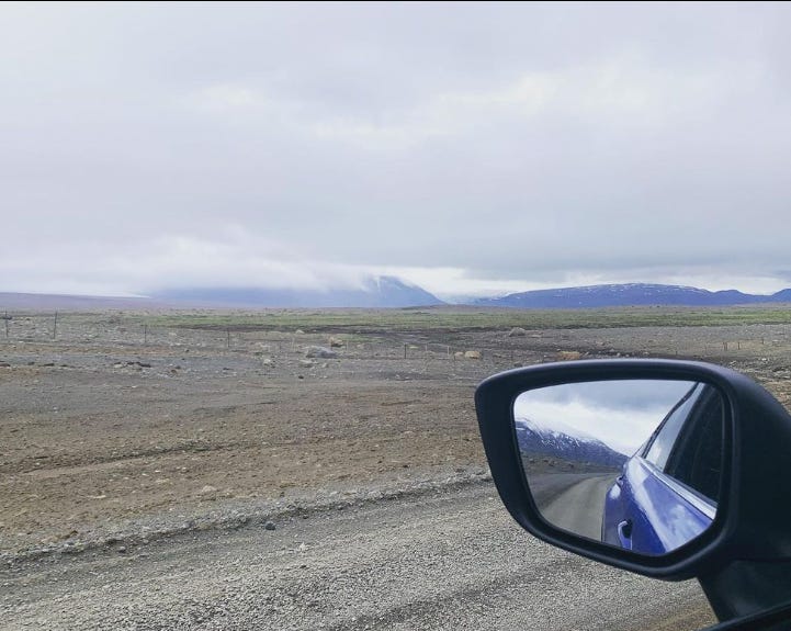 An image of rocky ground and blue mountains in the distance, taken out a car window with the rearview mirror also in the shot