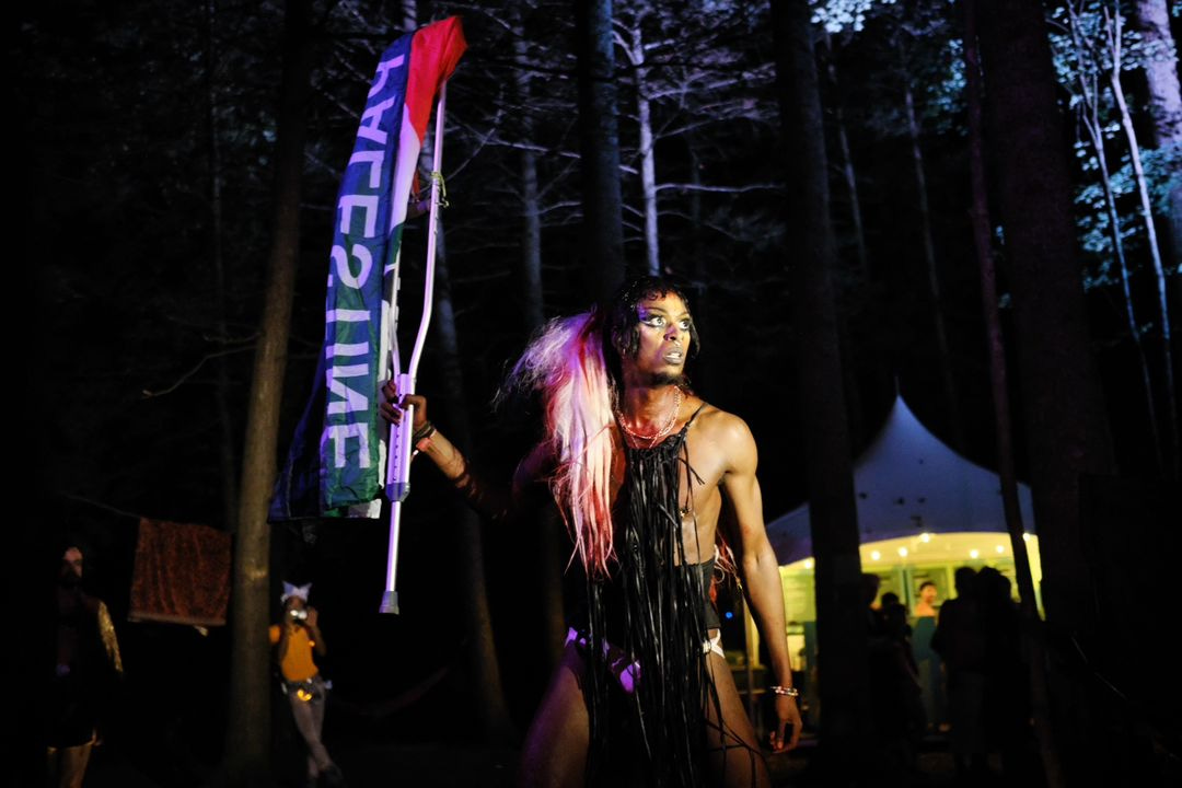 A Black performance artist is brightly lit in the woods at night. She’s holding an aluminum crutch with a flag that says PALESTINE. She is looking out in the distance, vibrant.