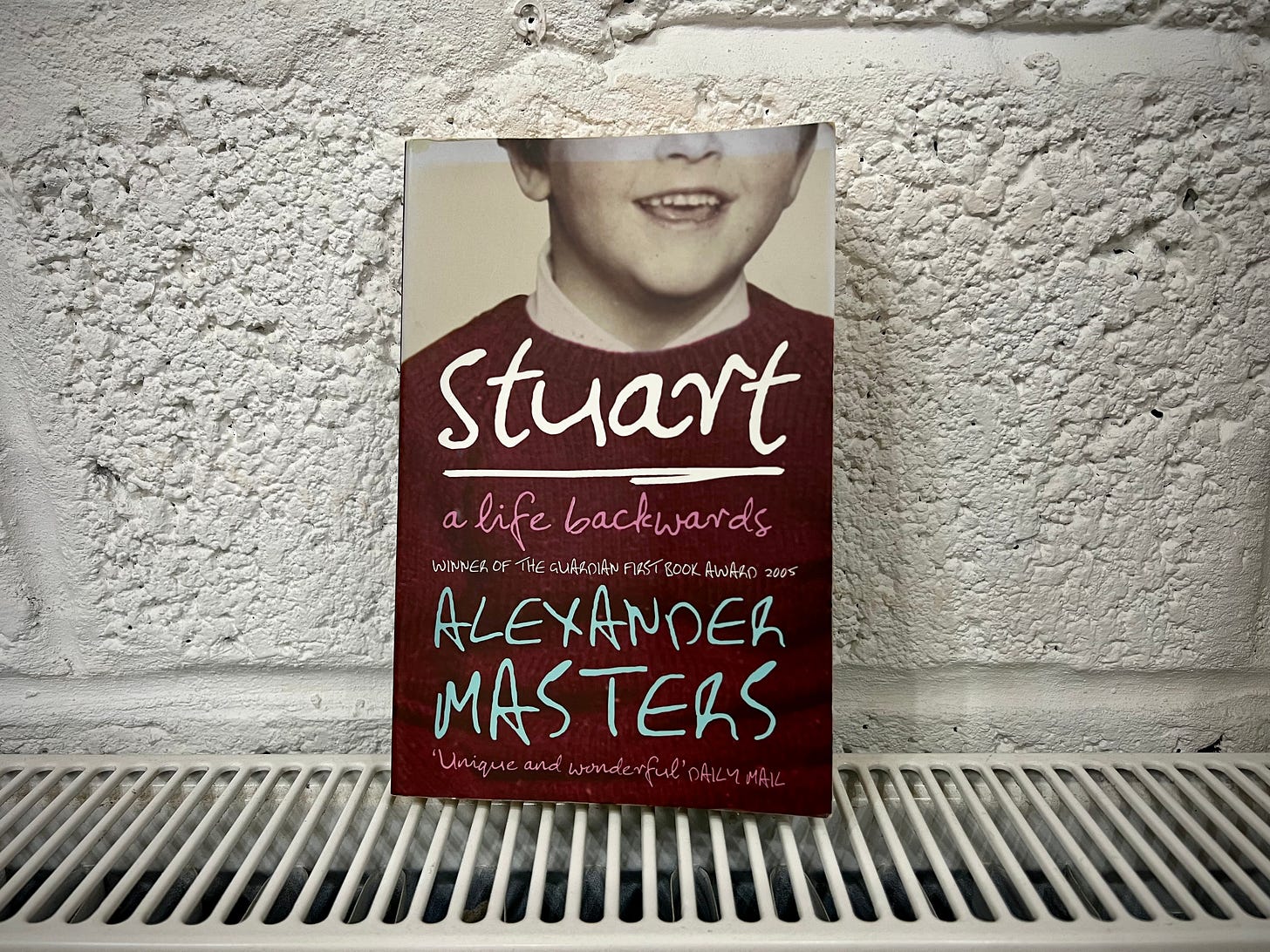 A photograph of the paperback novel Stewart, a life backwards. It's sitting on the radiator, leaning against a breeze, block wall, painted white.