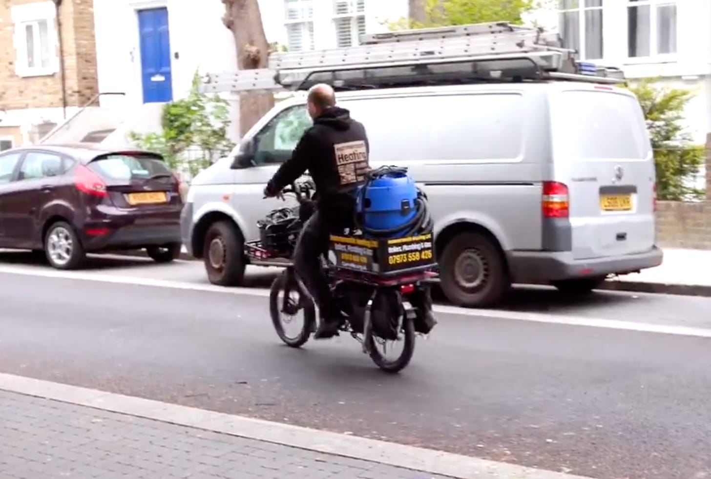 A man cycles away from the camera on a small electric cargobike. On the back of the bike is  perches a vaccum cleaner used for his work. There is signage on the side of the bike advertising his business. 