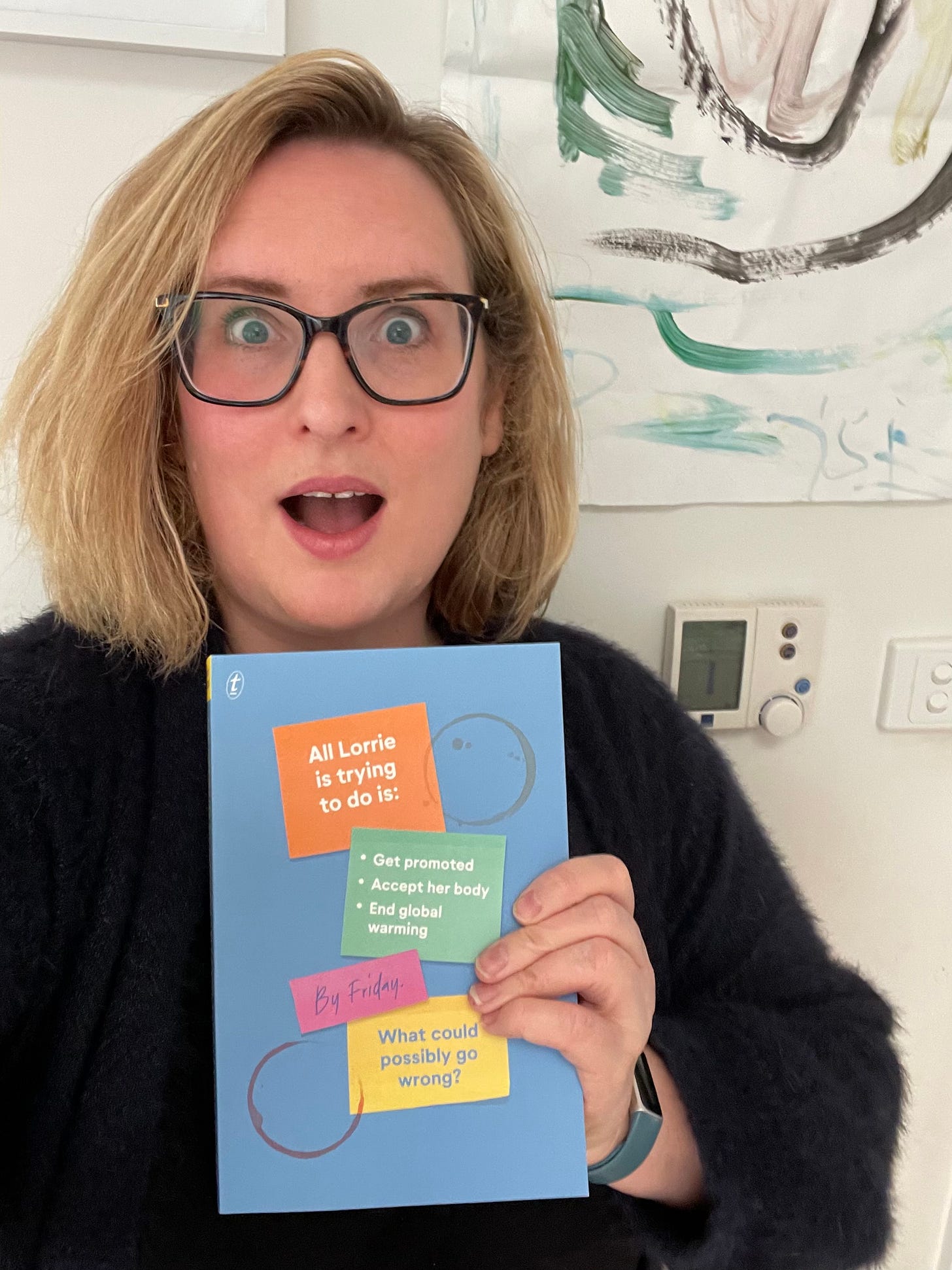 Photo of Eleanor, a woman in her forties with glasses and a messy blonde bob, making an exaggerated expression of shock/excitement while holding a copy of the proof of her novel. Cover is blue, with colourful sticky notes printed on the front with the text: "All Lorrie is trying to do is: get promoted, accept her body, end global warming - by Friday. What could possibly go wrong?"