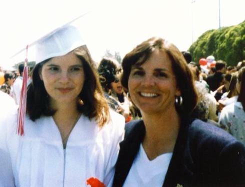 A photo of a high school girl in a white graduation cap and gown, with her mom at her side.