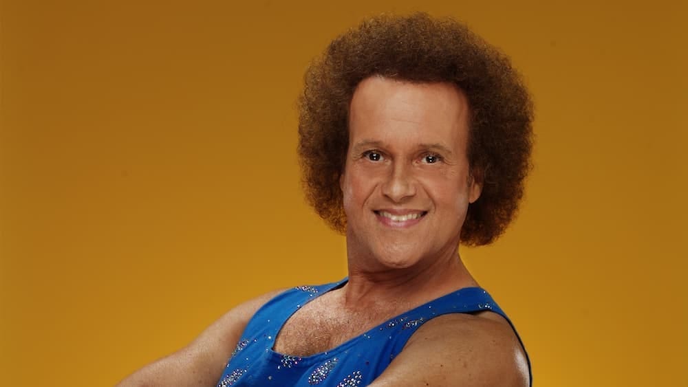 A photo of Richard Simmons wearing a blue tanktop and posing against a gold background