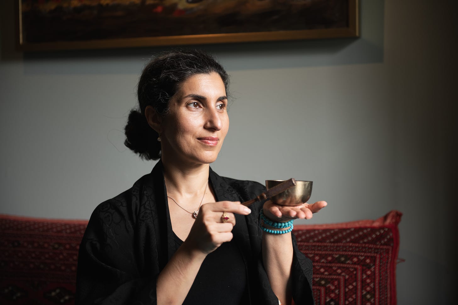 An Iranian-American woman with a gentle smile stares off to the right while gently tapping a small golden singing bowl in the palm of her hand.