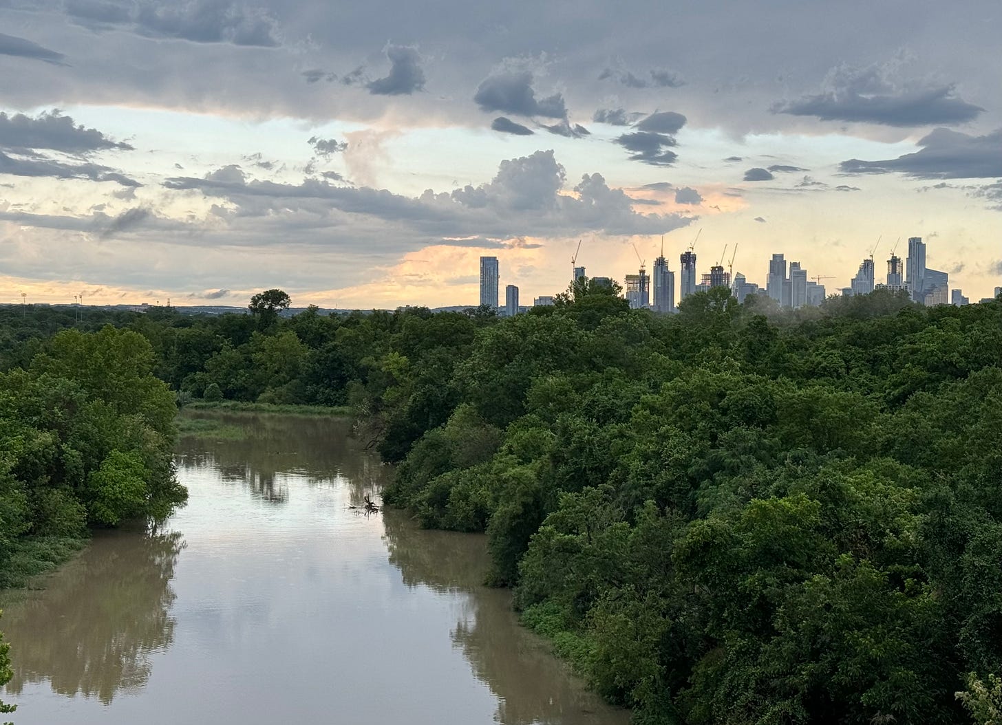 Colorado River and wild parklands; downtown Austin skyline with cranes visible in the background; sky clearing after a storm