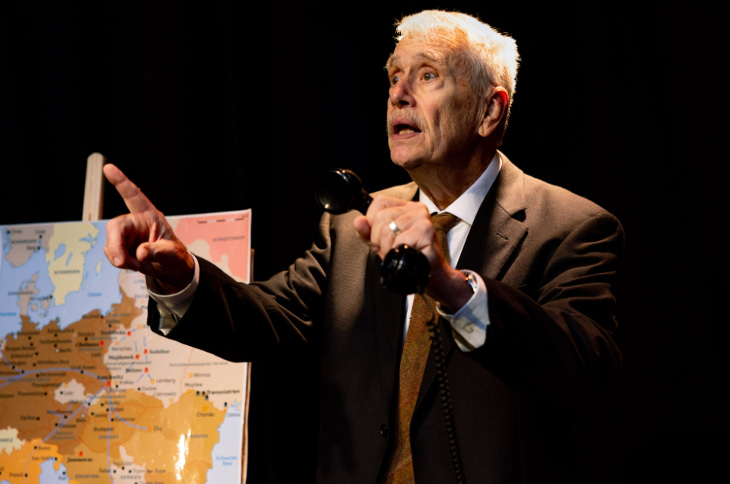 An actor on stage in a suit, in front of a large map of Europe