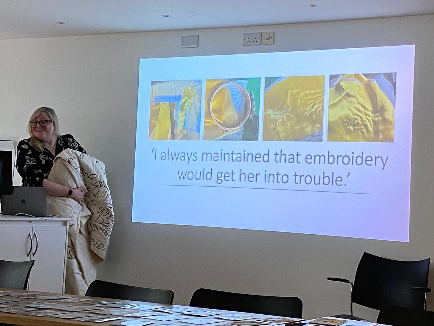 A white woman holding a large textile piece, folded, in front of a screen showing yellow silk and the words “I always maintained embroidery would get her into trouble”