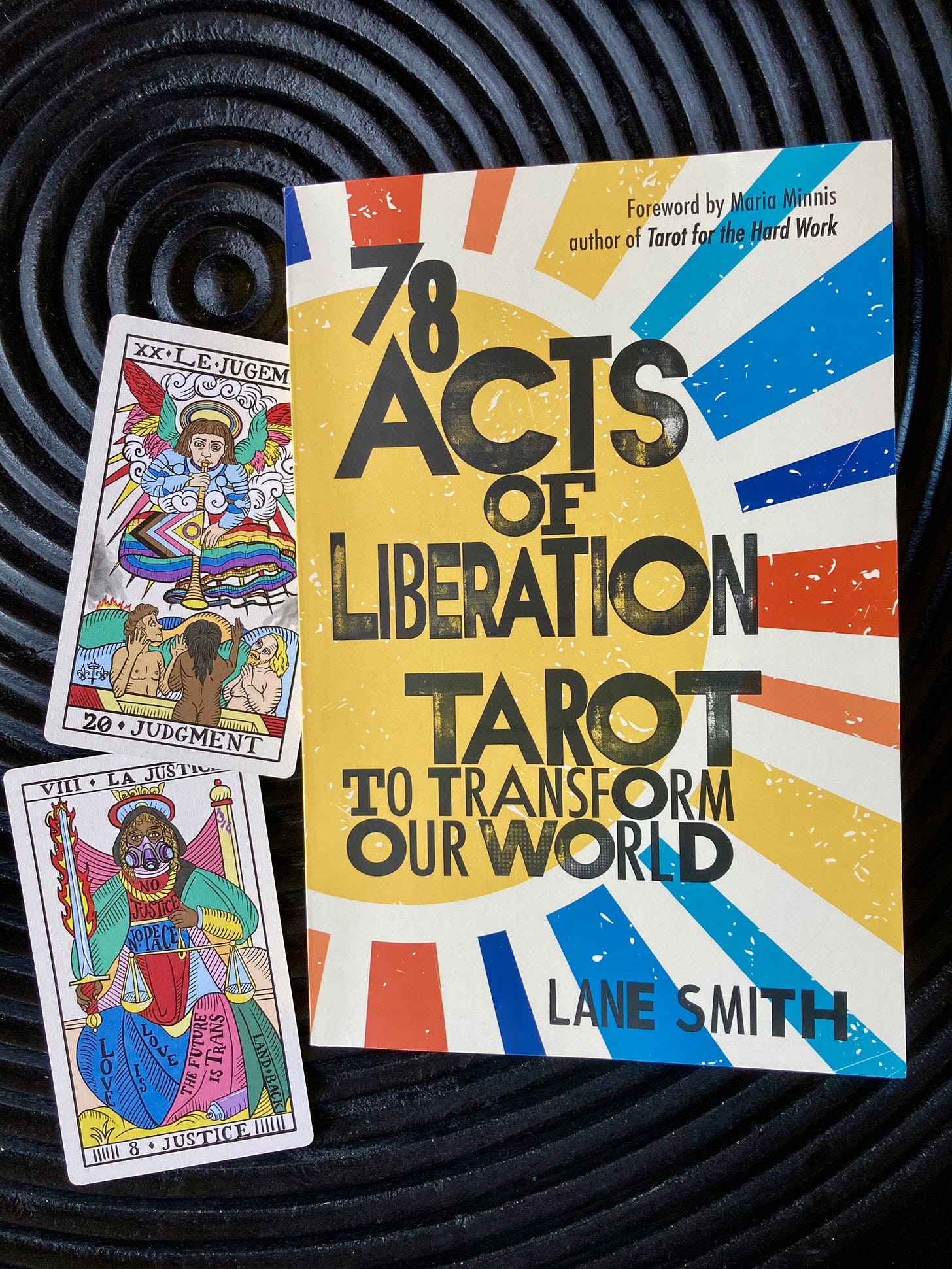 78 Acts of Liberation on a black surface next to the Judgment and Justice tarot cards. Judgment waves a Pride flag and Justice wears a gas mask with the BLM protest slogan, "No Justice, No Peace" emblazoned on their dress.