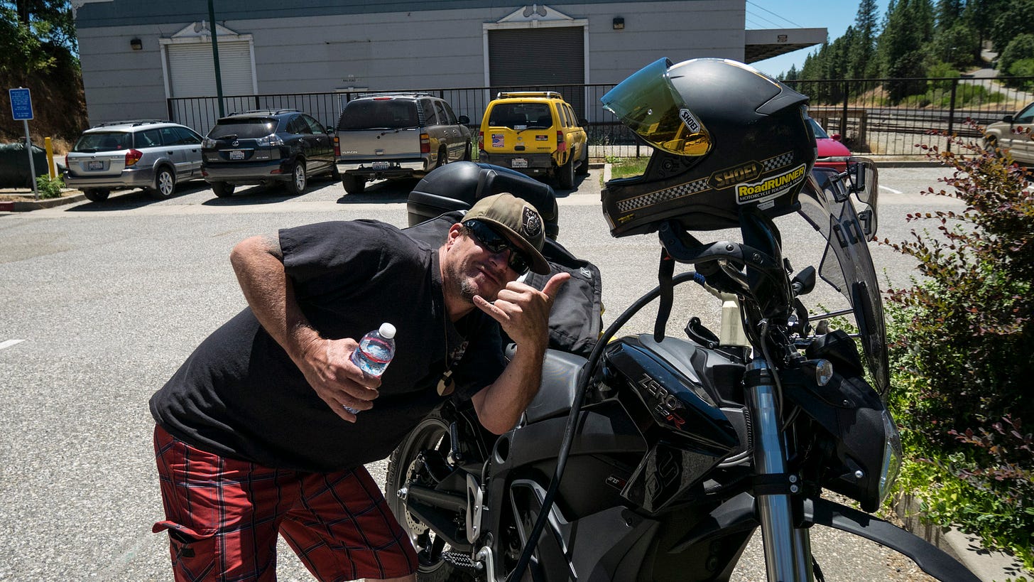 A man wearing red shorts, a black t-shirt, sunglasses, and an olive colored baseball cap crouches beside an electric motorcycle as it charges. The man is holding a bottle of water in his right hand and giving the "hook-em-horns" gesture with his left hand. He is leaning close to the bike and smiling. Beyond, a row of vehicles is parked beside a plain, gray building. To the right is some shrubbery in need of a trim.