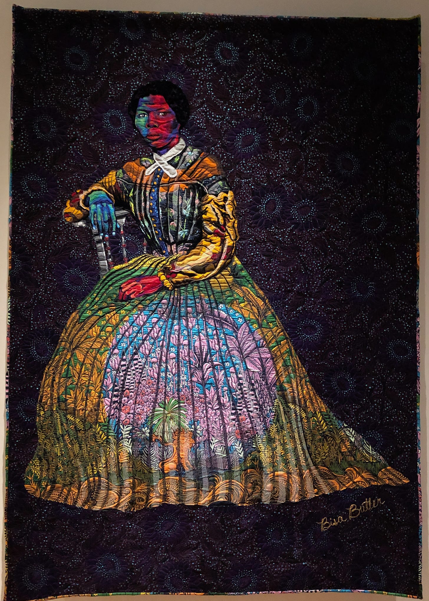 A multi-coloured fabric portrait of a woman sitting on a simple chair, at an angle, her right arm resting on the back of the chair. She wears a long-sleeved dress with a full skirt, and a cravat. The words “Bisa Butler” are written at an angle on the bottom right-hand corner.
