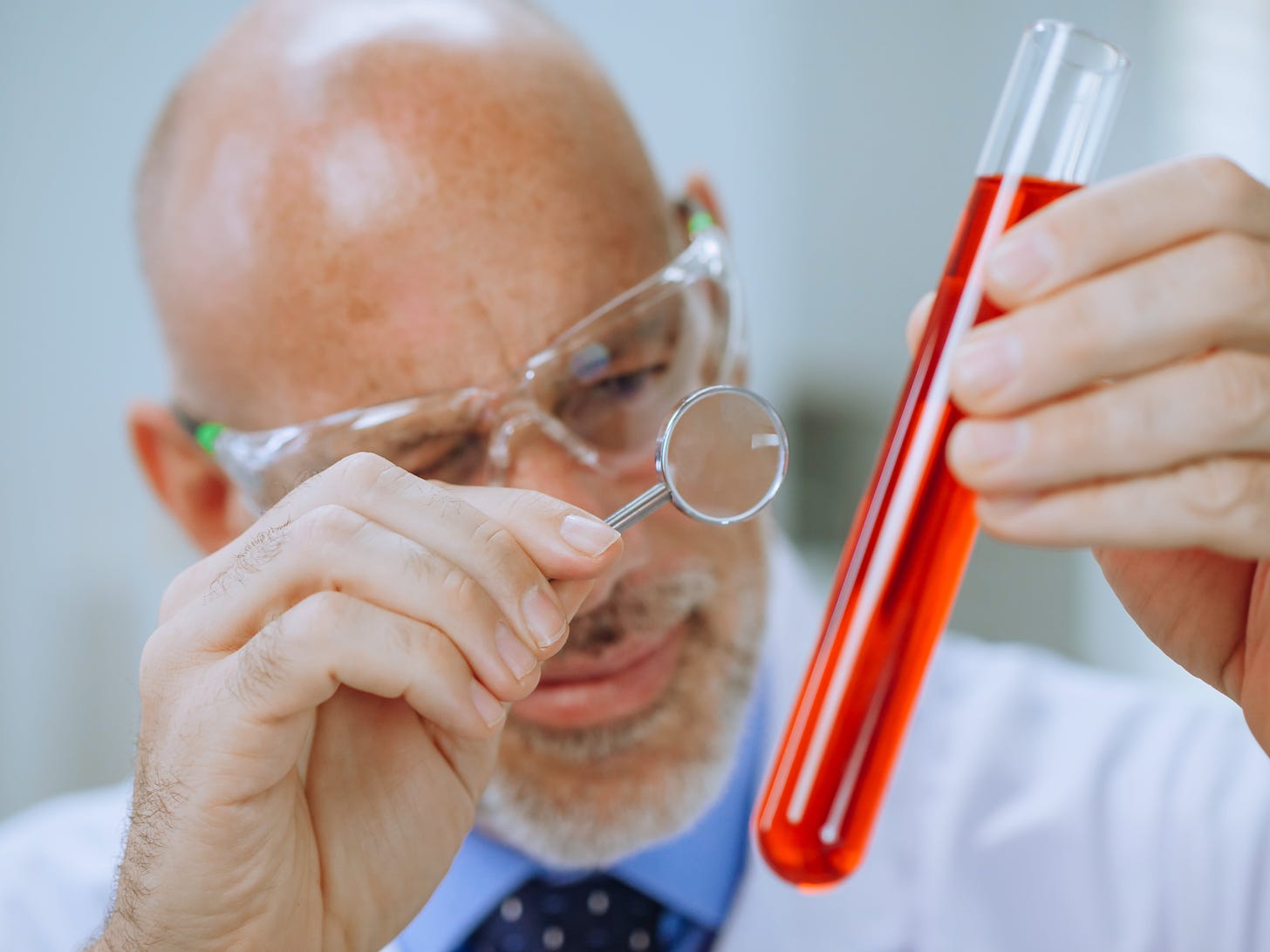 Photo of an older man in a lab coat examining a test tube filled with red liquid using a magnifying glass. That is to say, there is no actual science taking place in the photo, just the veneer of "science-ness." 