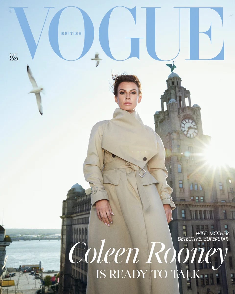 Image shows Coleen Rooney standing in front of the Liver Building in Liverpool, with the River Mersey visible in the background. She is gazing into the camera wearing a beige belted trench coat, and her brown hair is swept back into an updo. The breeze is whipping her hair around her face and seagulls circle overhead. Above her head, the British Vogue logo in blue. The cover line reads: “Wife, mother, detective, superstar. Coleen Rooney is ready to talk.”