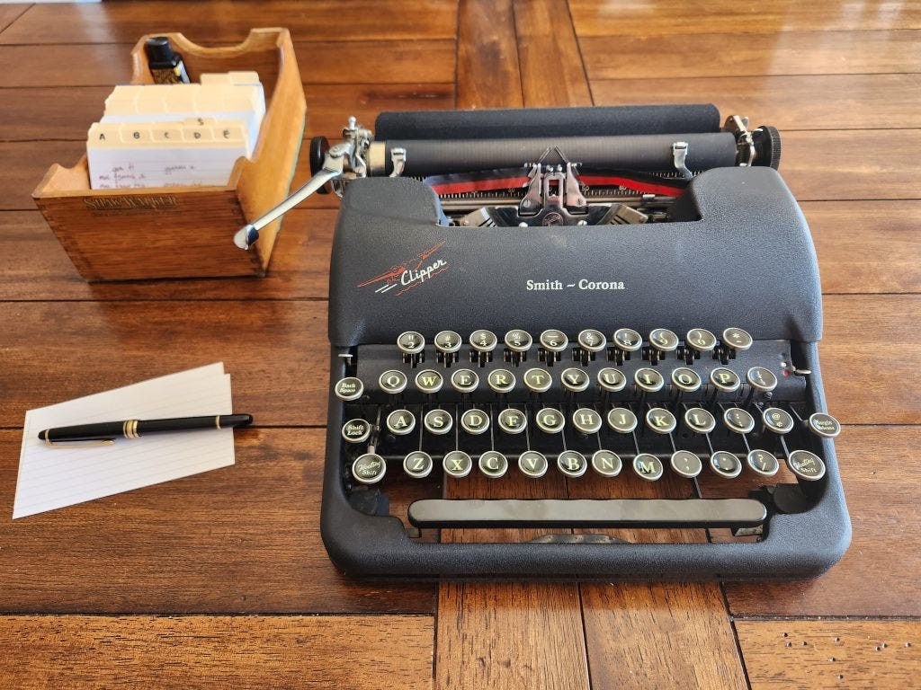 A wooden table arranged with a black Smith-Corona Clipper typewriter next to a Shaw-Walker wooden dovetailed card index, some index cards, and a black fountain pen.