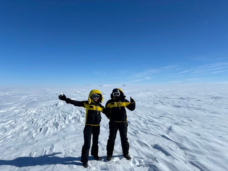 Two people in winter gear stand on a field of white ice with blue sky above them. Two people in winter gear stand on a field of white ice with blue sky above them.