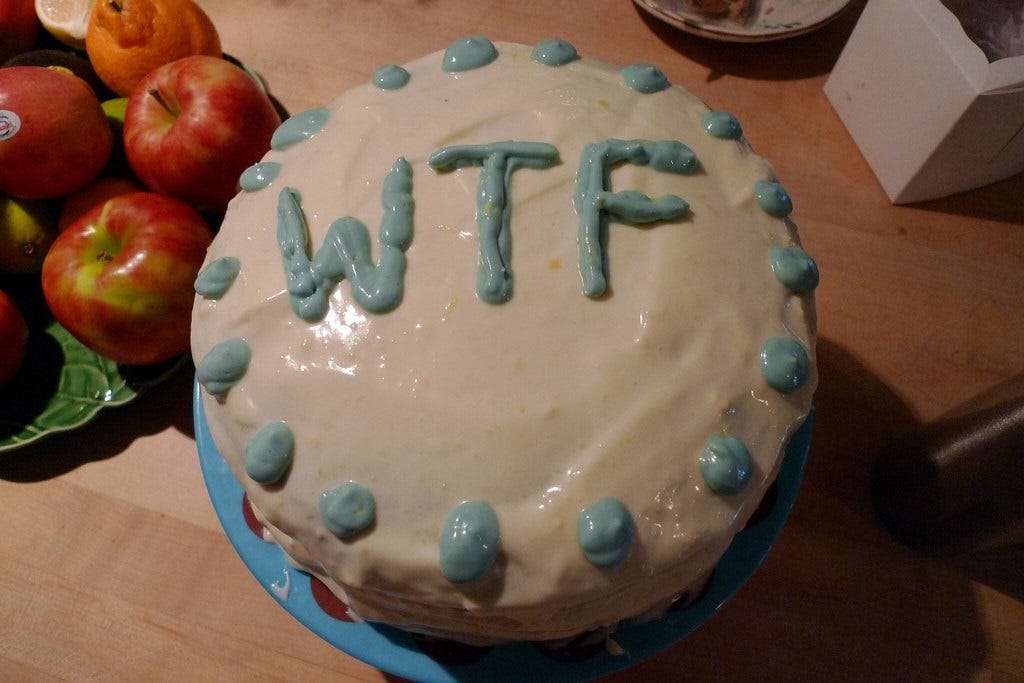 Photo of a round layer cake on a blue plate on a table. The cake's white icing is decorated with blue icing dots around the top, and large letters in blue icing reading 'WTF' (no question mark). To the upper left of the cake is a tray of apples. Photo of a round layer cake on a blue plate on a table. The cake's white icing is decorated with blue icing dots around the top, and large letters in blue icing reading 'WTF' (no question mark). To the upper left of the cake is a tray of apples.