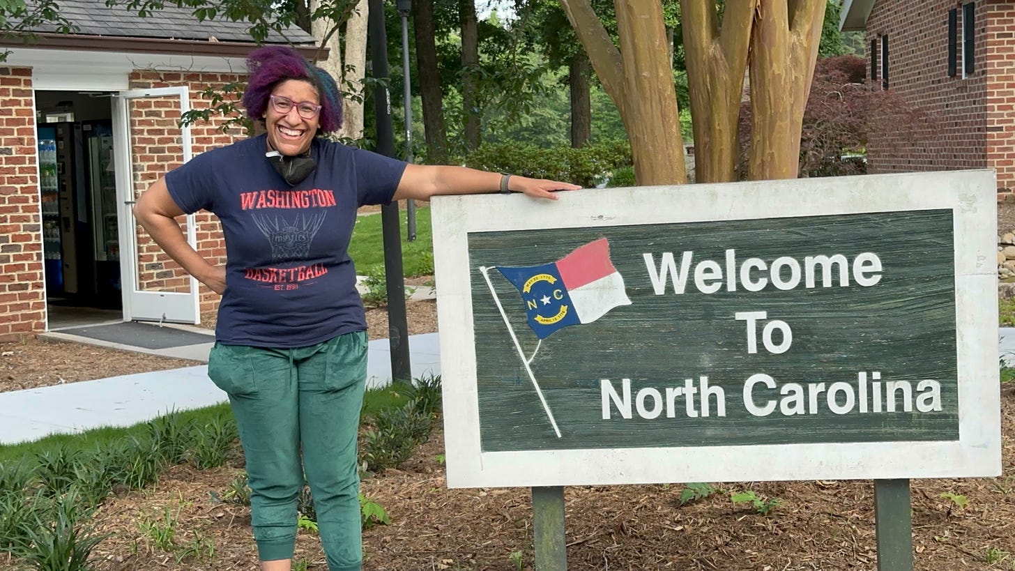 Kristen is wearing green sweats, a blue t-shirt and has her mask off briefly to make a video as she stands next to the welcome to north carolina sign