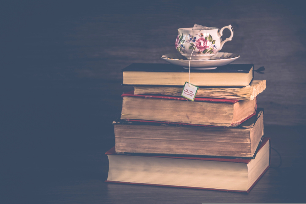 A stack of old books, their spines facing away from the camera, topped by a flowered teacup with a teabag tag and string hanging over the side