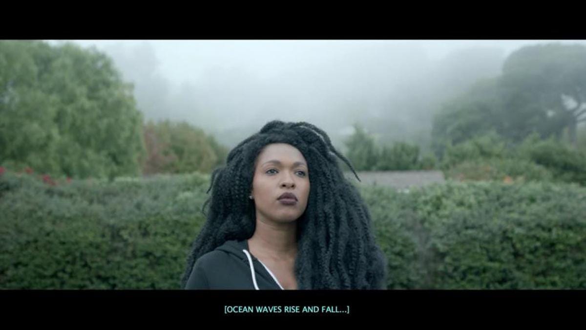 A still from The Tuba Thieves. A Black woman in a hoodie stands in front of a garden hedge. A blue caption reads [OCEAN WAVES RISE AND FALL…]