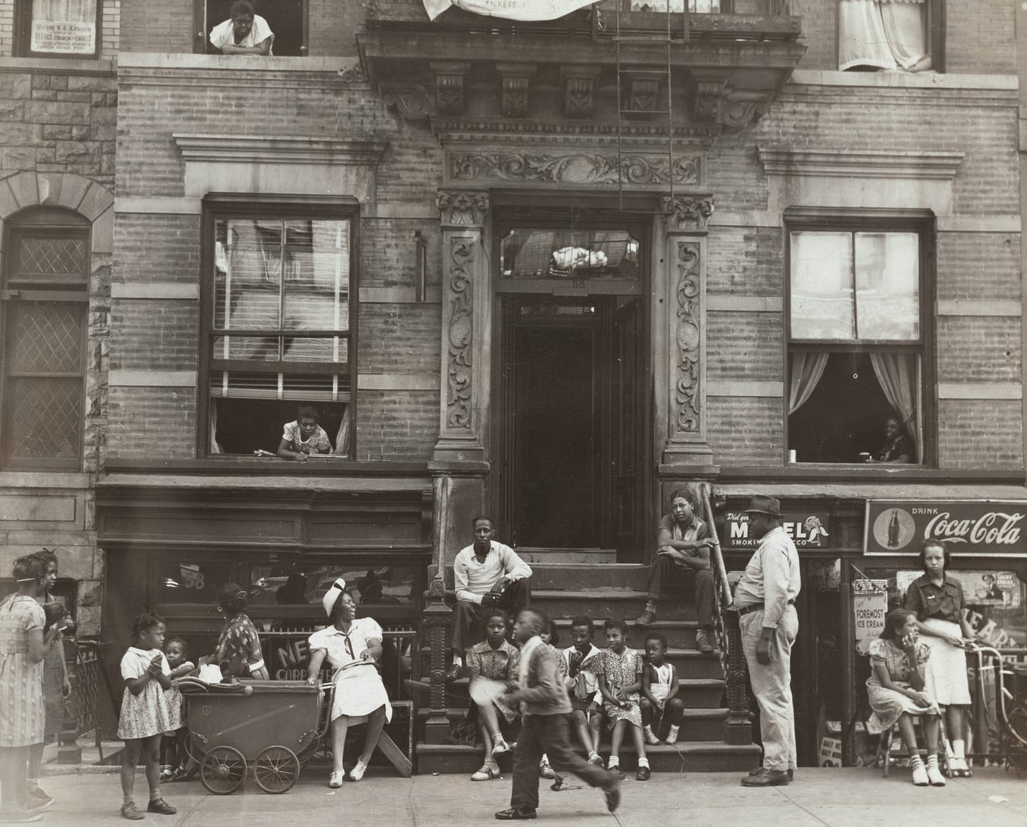 Black and White image of brick apartment building in early 20th century with Black children and parents sitting on steps and playing on the sidewalk