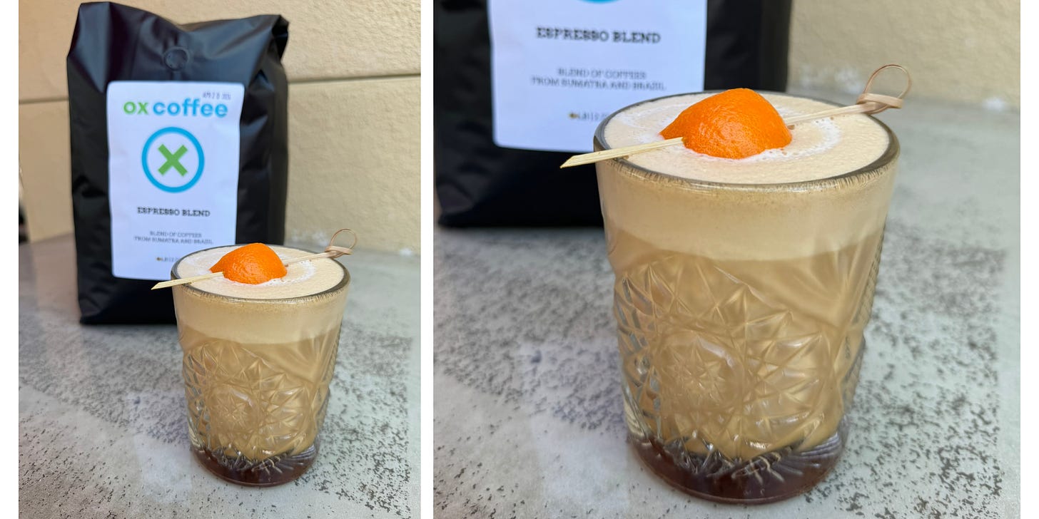 A close-up of a coffee drink with an orange peel resting in a thick layer of foam in an clear glass tumbler.