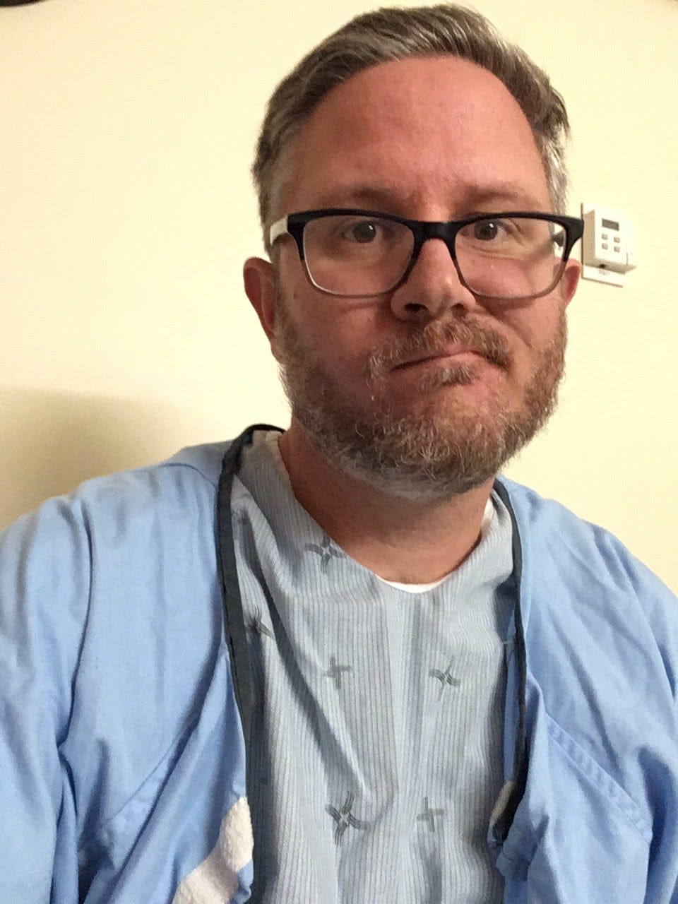A selfie of me from the very next day. I am wearing a johnny shirt at the hospital. I look unhappy.