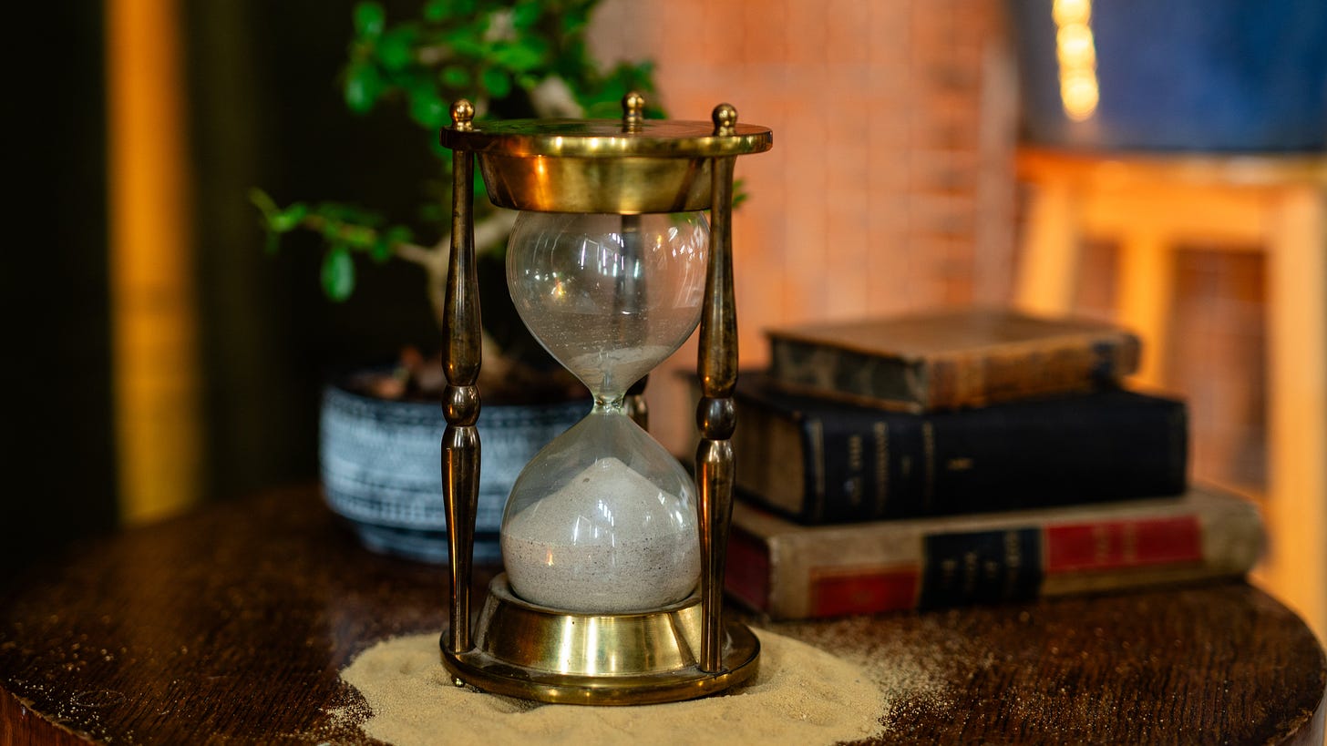 An hourglass sits on a dark wooden coffee table in front of some old books and a bonsai tree in a pot.