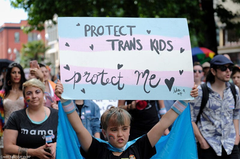 A child holds a blue, white, and pink sign saying “protect trans kids, protect me” as they stand among a crowd of pride parade attendees