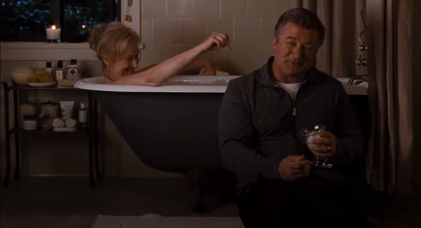Movie still from It's Complicated. A woman lounger in a bathtub while a man sits outside of it, eating ice cream in a glass.