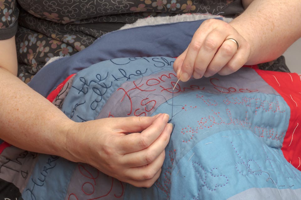 Two hands, one holding a needle, the other holding thread, stitch on a quilt.