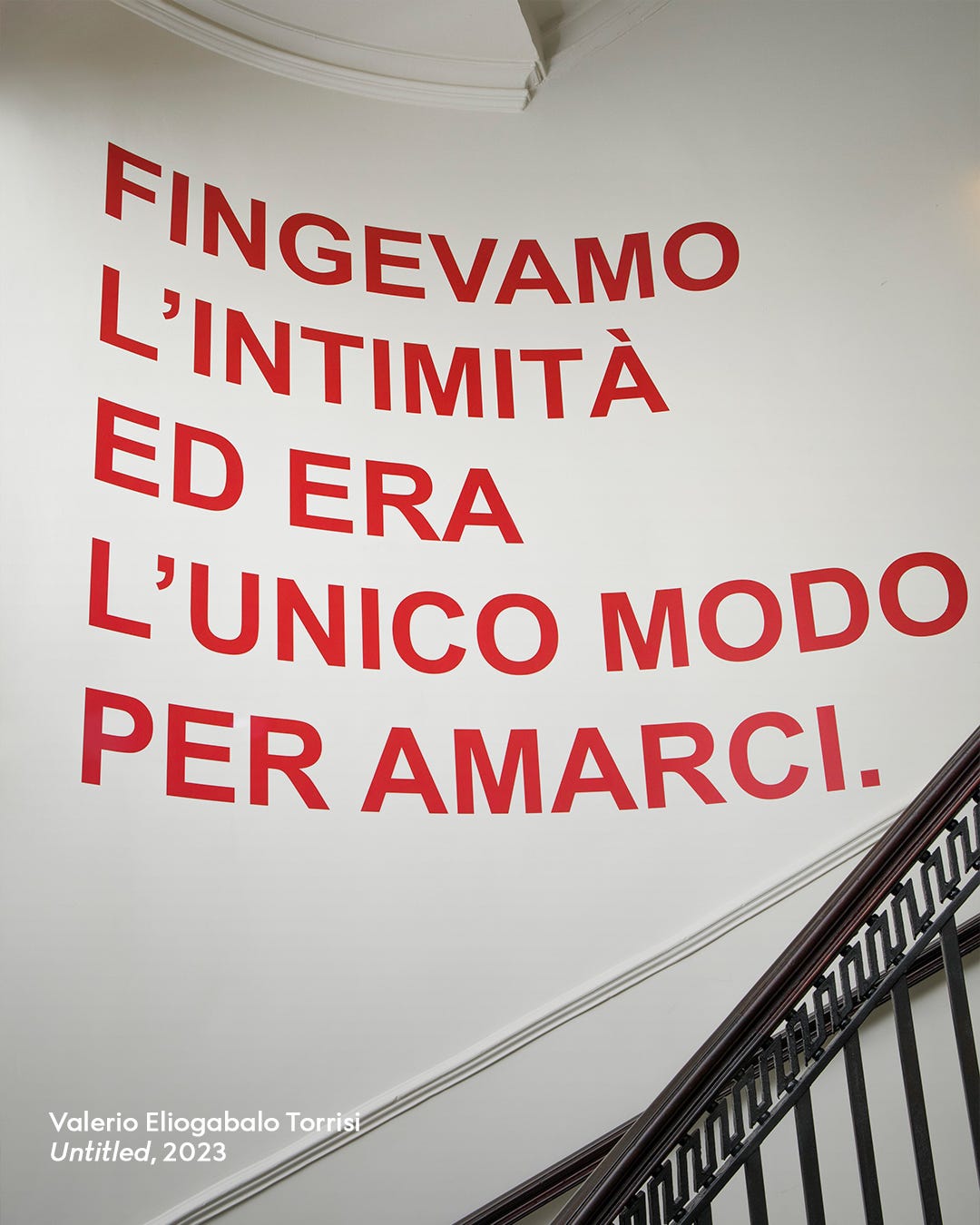 "Fingevamo l'intimità ed era l'unica modo per amarci" is written in red on a white wall in front of an iron spiral staircase. 