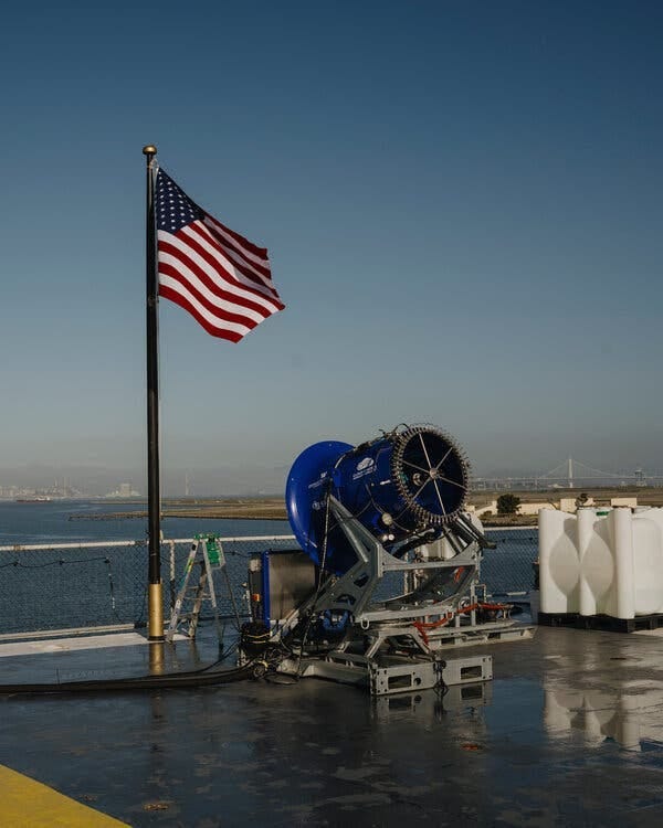 A view of the spraying machine, looking something like a short cannon. The barrel is royal blue. A United States flag waves on a short mast just behind, at the edge of the carrier’s flight deck. 