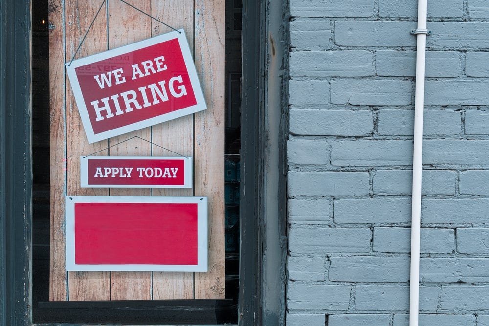 A couple of signs hanging on a shop window. “We are hiring,” and “Apply today!” A couple of signs hanging on a shop window. “We are hiring,” and “Apply today!”