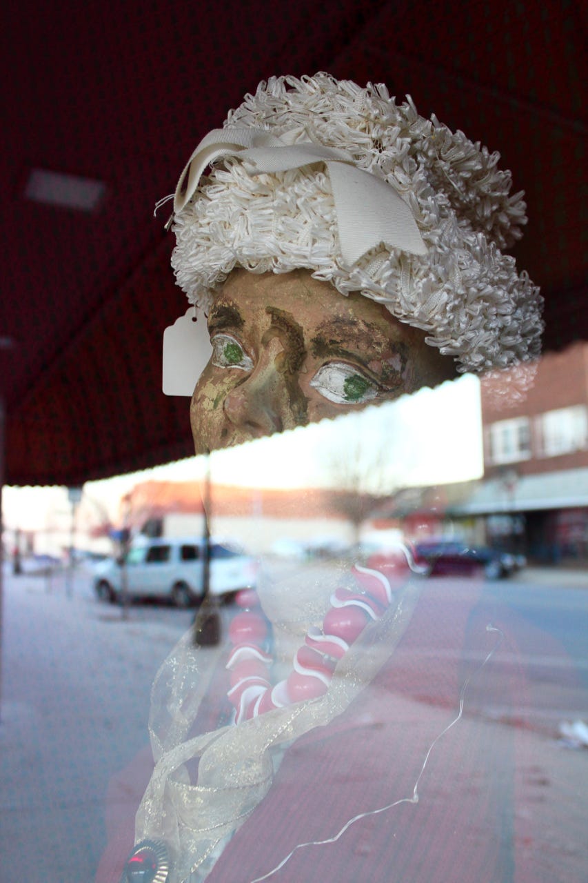a color photo of a vintage manikin wearing a vintage hat, both items being displayed in a shopfront window on a small town's street. half the photo is a reflection on the glass window of the street scene itself. 