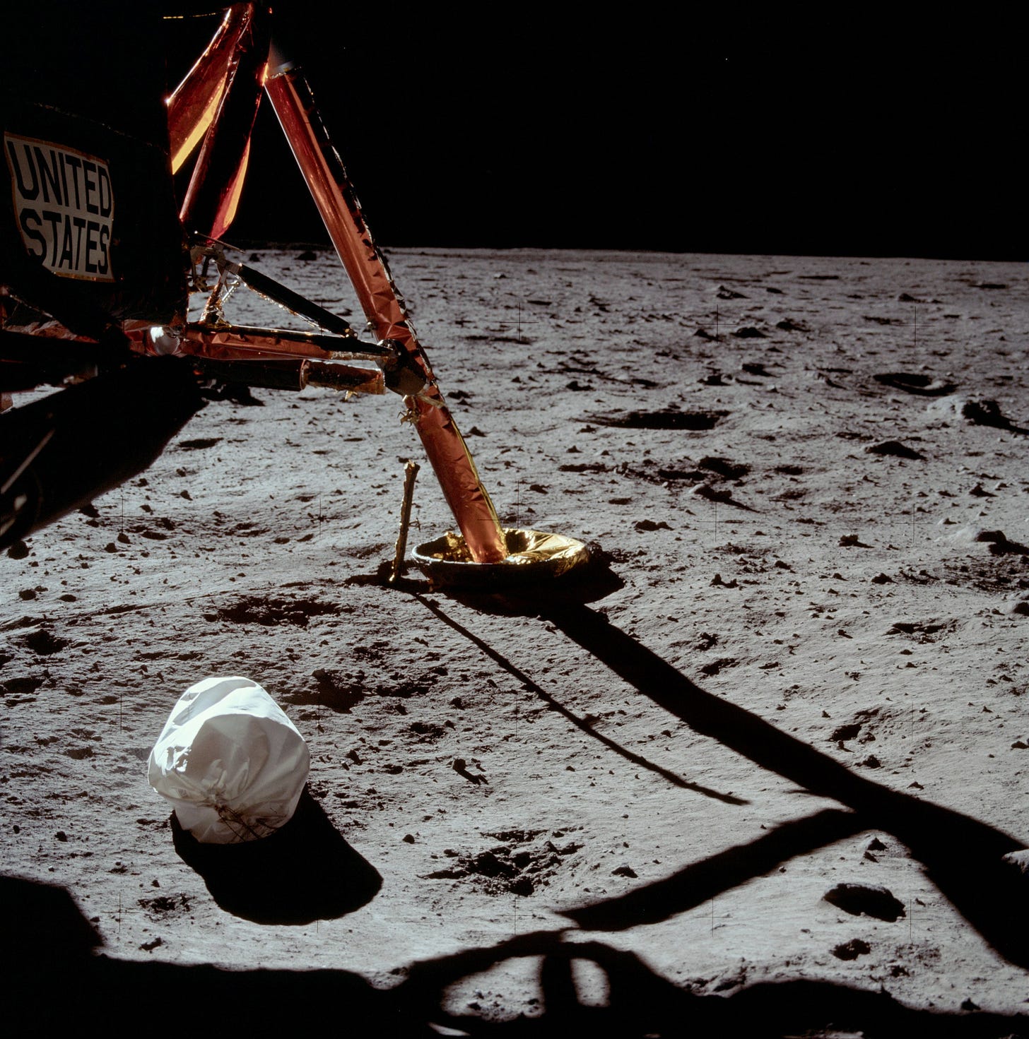 A color photo taken on the Moon's surface. The horizon is in the distance, with an empty black sky above. In the foreground is the landing strut of the Descent Stage, with "United States" written on the side of the craft. A white fabric bag lies underneath the craft.