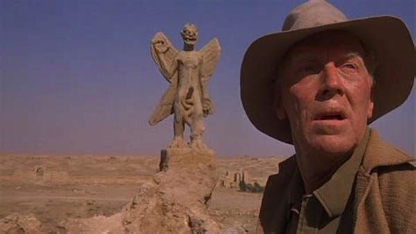 An ancient stone statue of a demon, standing atop a worn sandstone pedastal. In the foreground an older man in dusty clothing on an archeologist, looks away from the statue in horror. Pazuzu is often depicted as a combination of animal and human parts with his right hand pointing upwards and his left hand downwards. He has the body of a man, the head of a lion or dog, eagle-like taloned feet, two pairs of wings, a scorpion's tail, and a serpentine penis.