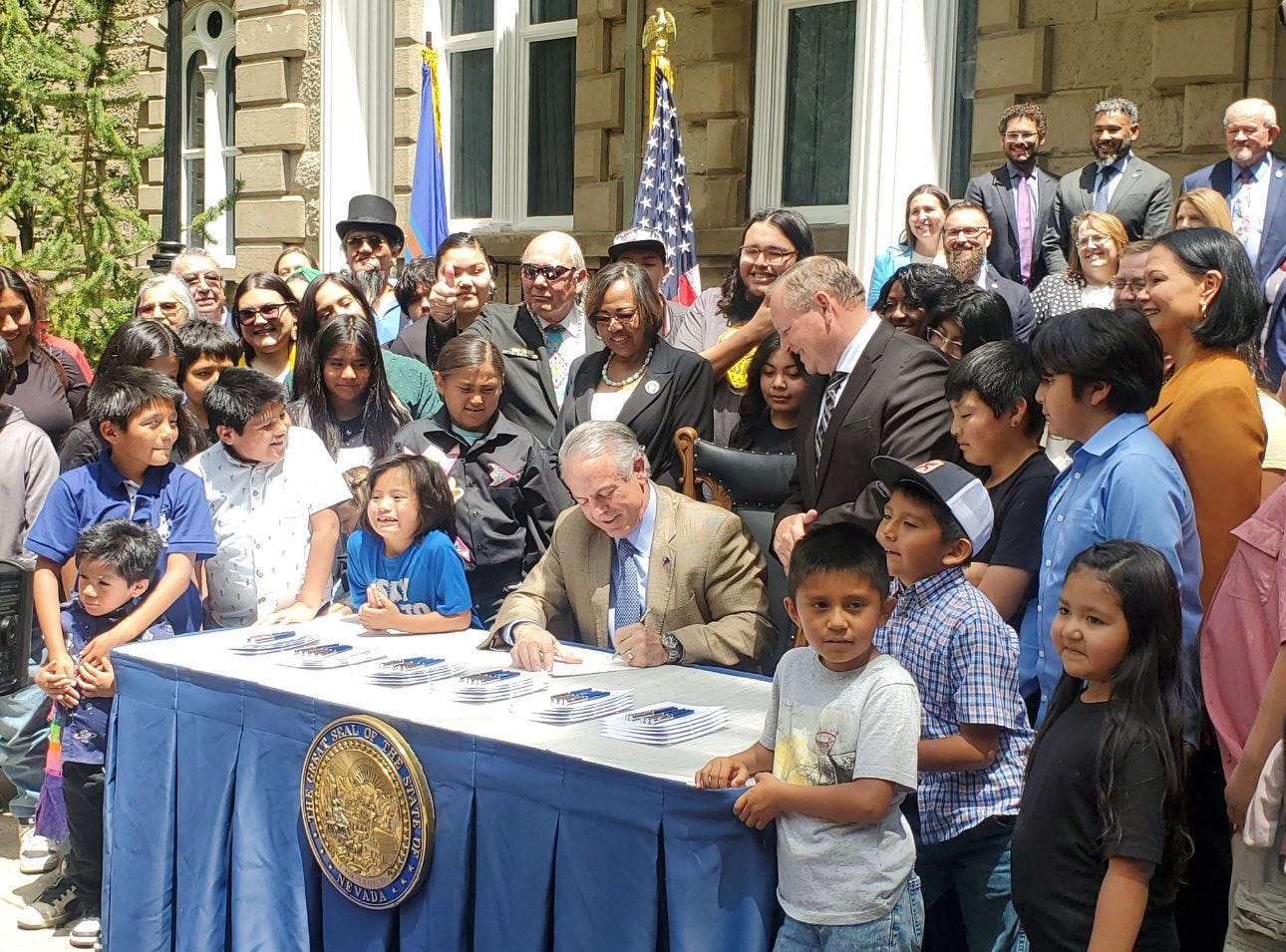 Nevada Governor Joe Lombardo seated at table signing Assembly Bill 519. He is surrounded by children and tribal leaders from the Duck Valley Reservation and Nevada Legislative members.