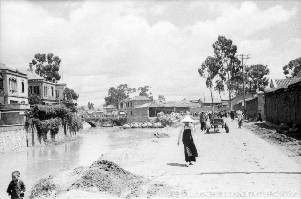 A black and white photo of a canal in the city of Hanoi as seen in the fall of 1940. A woman walks down a dirt path holding a conical hat while carts and people travel the road behind her.