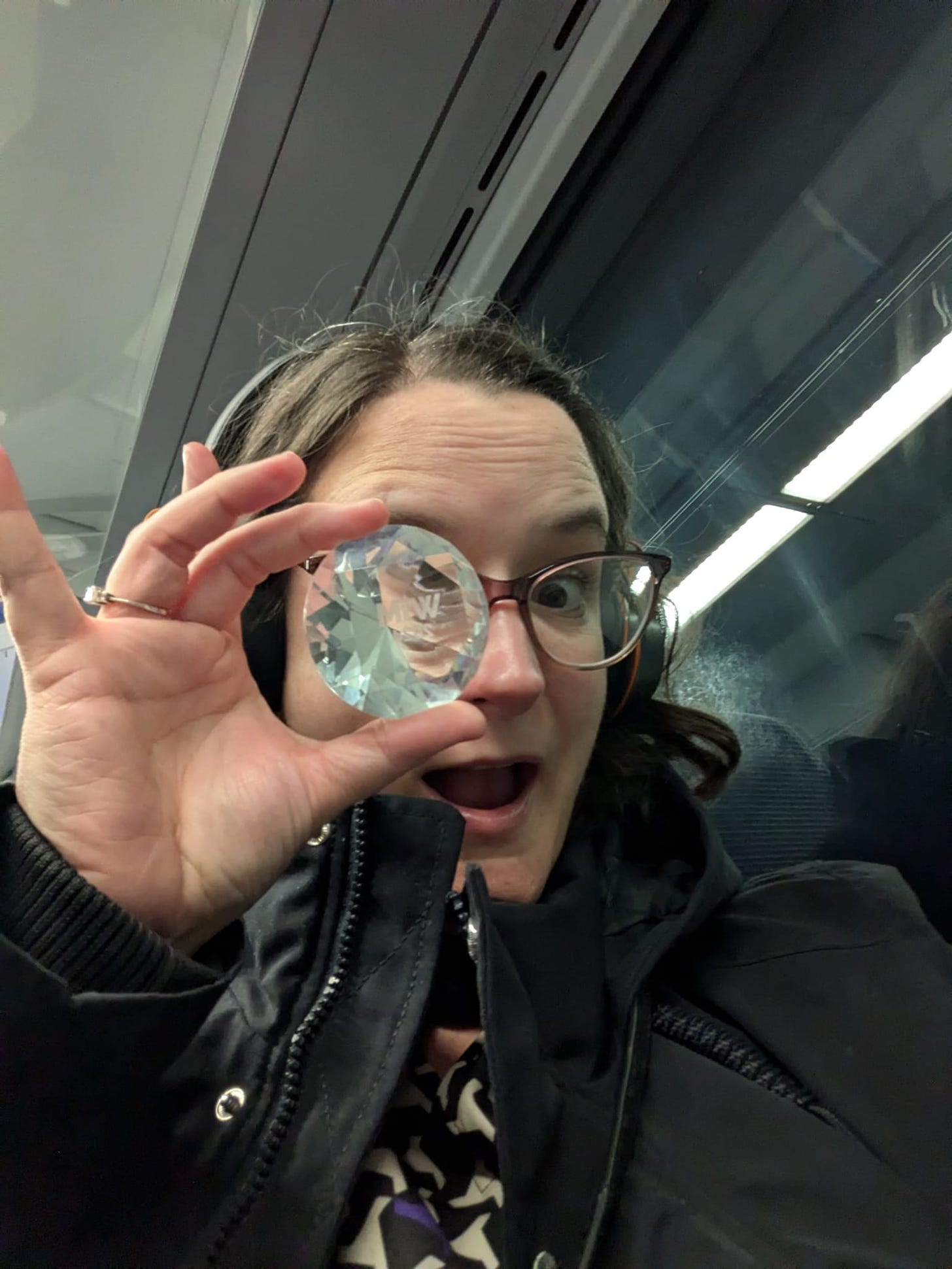 Mel Barfield, a woman in her 40s with brown hair wearing over-ear headphones and glasses is bundled up in a big coat while sitting on a night time train. Her right eye is obscured as she's holding up the small crystal trophy she won at the Women In Marketing Awards.