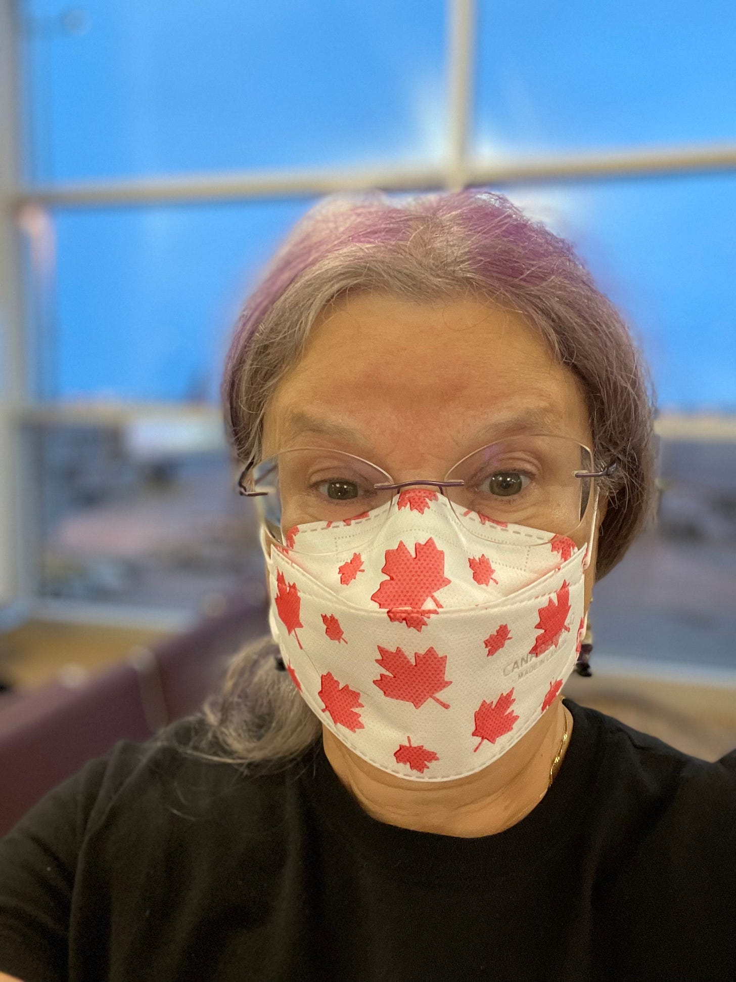Silver and purple haired femme with glasses wearing a white KN95 mask with red maple leaf images on it, with a blue sky through a window in the background