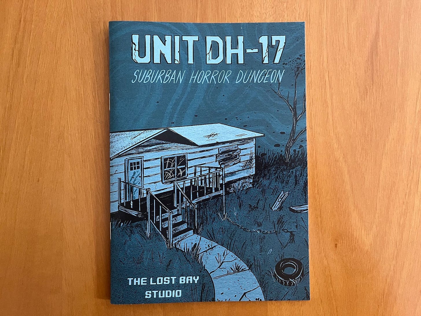 The zine Unit DH-17, designed by The Lost Bay Studio, written for The Lost Bay. The cover is entirely shades of blue, and features a dilapidated trailer home. The lawn is overgrown and littered with scrap wood, as well as a tire and a chain. One window is cracked and the other is boarded up. A spindly and leaf-less tree can be seen in the background.