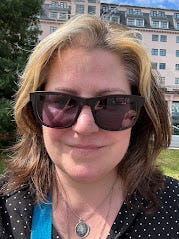 A close up go my head and shoulders, standing outside, with a pale building in the background. I've got a blonde fringe and brown hair, I'm wearing a black top with small white spots, black sunglasses in a rayban style, and have a silver compass pendant round my neck. My smile is visible.