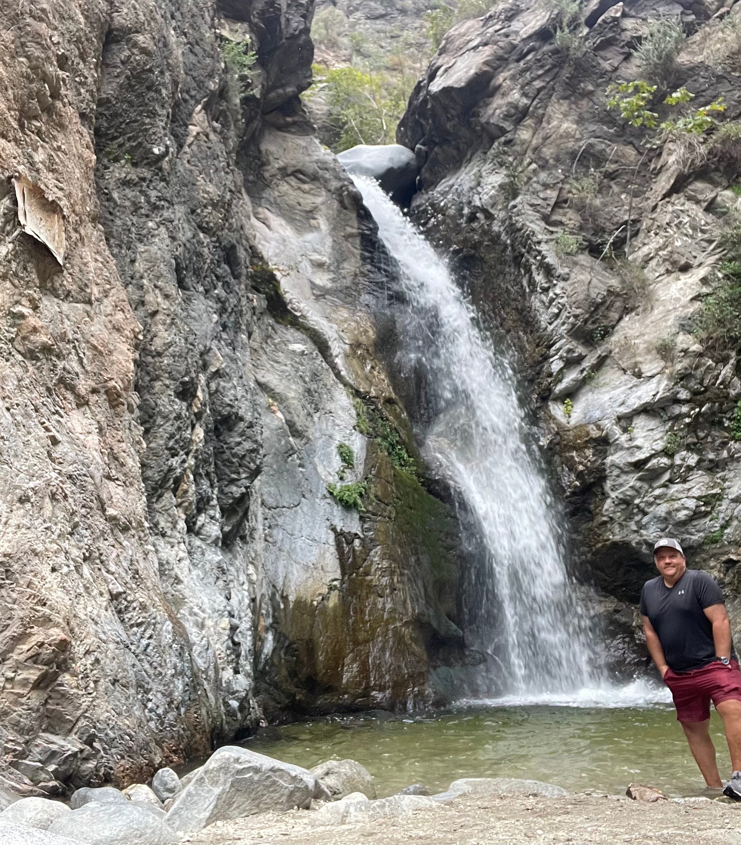 A small waterfall cascading dow into a pool between two rock walls, mickey is to the right, wearing maroon shorts and a black t-shirt