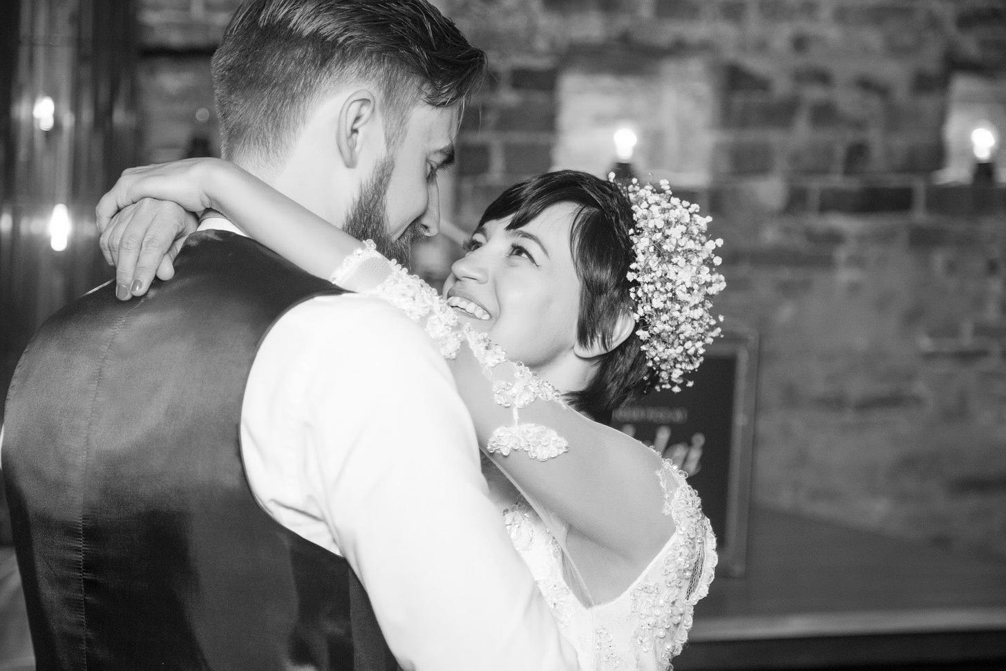Imagem preta e branco: Casal vestidos de noivos abraçados se olhando. Os dois estão sorrindo. Ela tem cabelo curto e um casquete de flores na cabeça. 