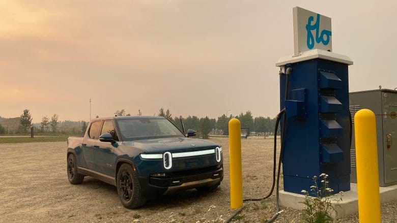 A blue electric truck is parked next to a charging station against a backdrop of a smoke-filled, orange sky.