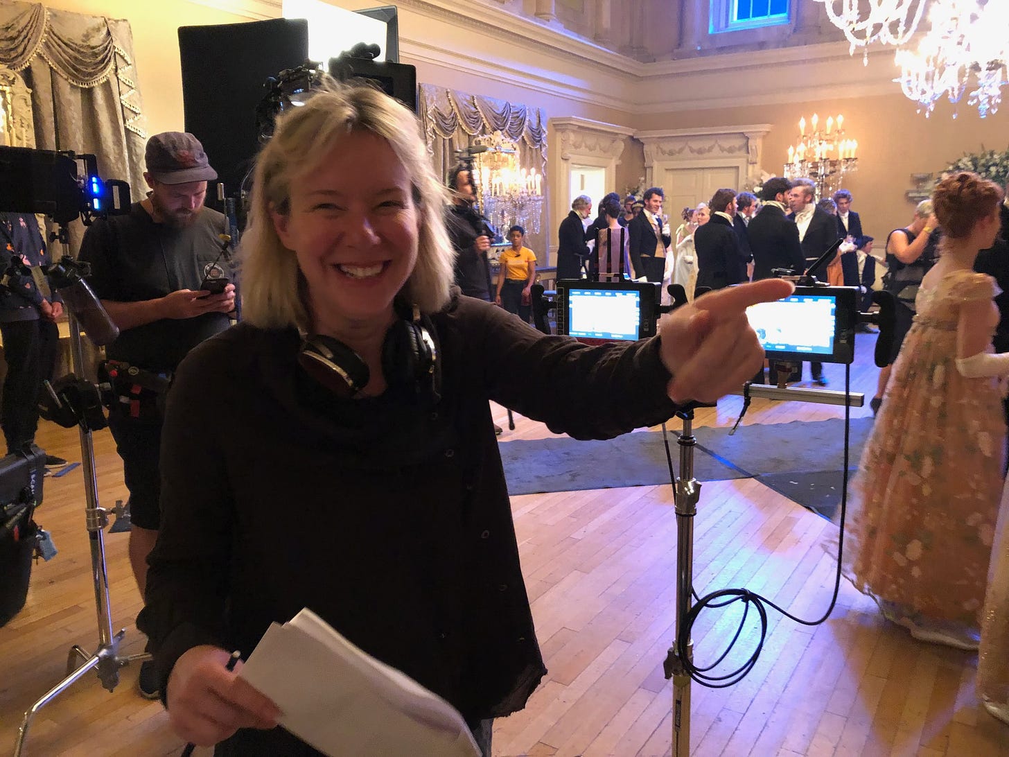 Film director Julie Anne Robinson on the set of Bridgerton. Behind her are actors in period costume, inside a ball room. You can see the film crew, lights, and cameras in the foreground.