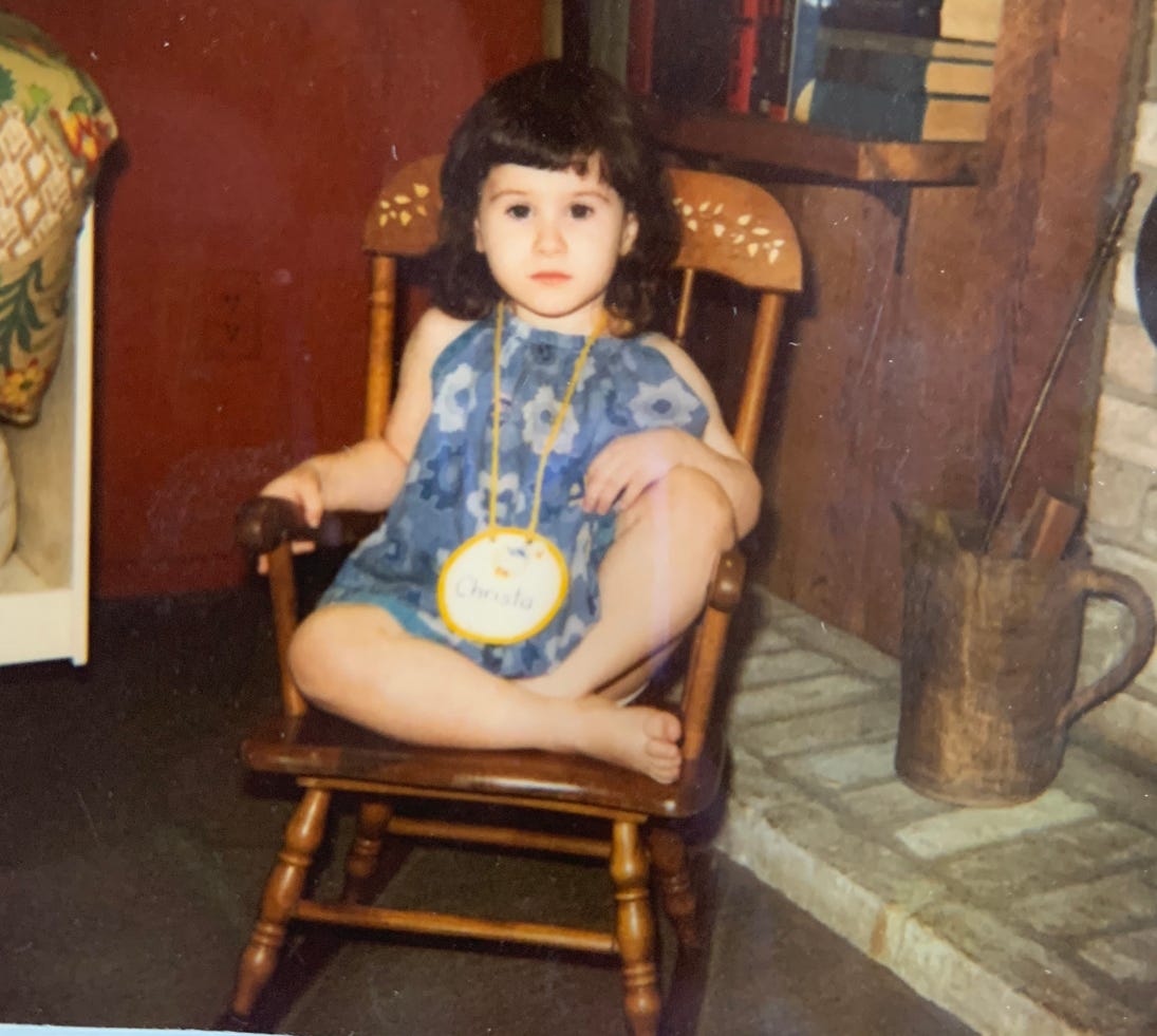 Photo of a serious looking child with dark hair and fair skin in a blue dress. They sit in the rocking chair blank faced and sullen.