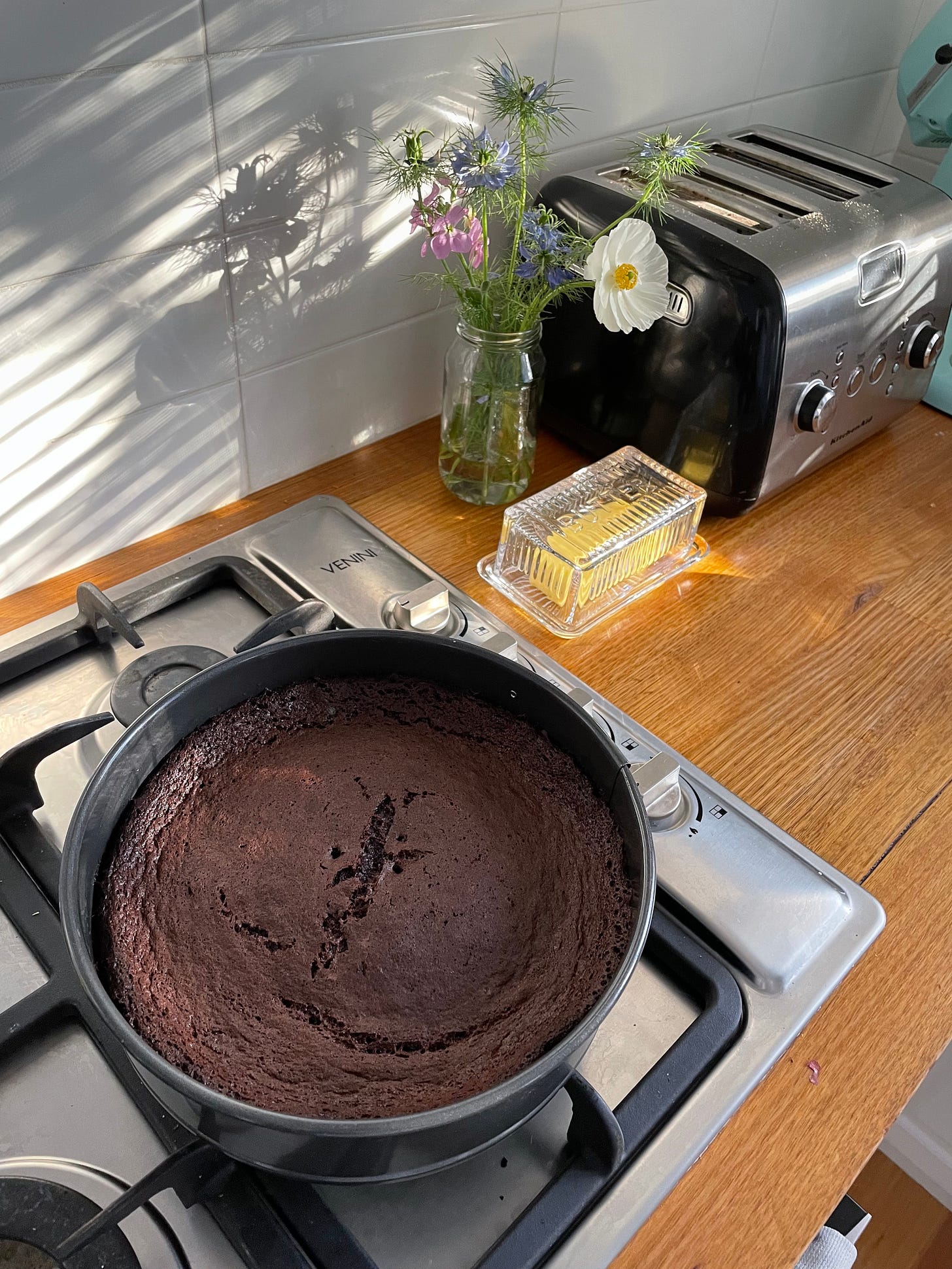 Dark chocolate cake cooling on the stove in a spring form tin. It's lit by sunlight, a posy of flowers, toaster and butter dish on the kitchen counter.