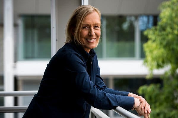 The tennis star Martina Navratilova smiles in a portrait photograph from 2021. She stands on a balcony, leaning on the railing.