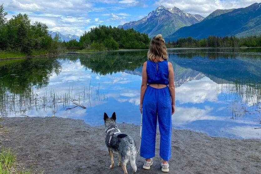 Scout the blue heeler stands on the edge of a large lake next to her owner Haley, a young woman in a blue jumpsuit. There are mountains in the background reflecting on the water's surface.